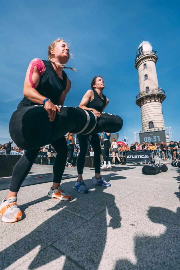 Zwei Athletinnen beim Partner-Carry vor dem Warnemünder Leuchtturm