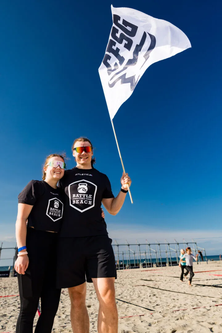 Zwei Crew-Mitglieder mit Battle-the-Beach-Flagge am Strand